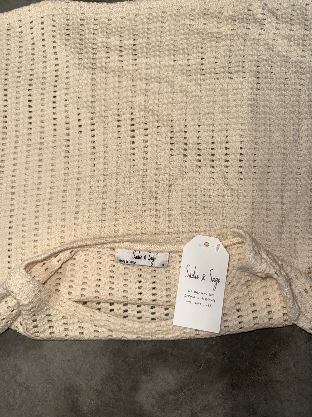 A close-up of a cream-colored, open-knit crochet fabric accessory, likely a bag or pouch, featuring a drawstring closure and a white paper tag with handwritten details.