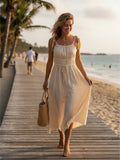 Yanet Collection Shop beige midi sundress with tie straps and gathered waist, worn by a woman walking on a beach path