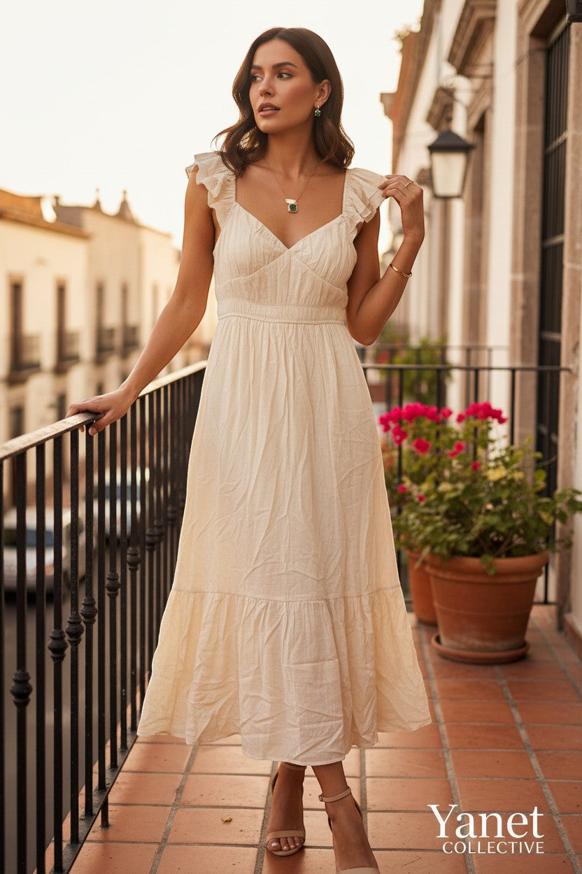 Yanet Collection Shop cream maxi dress with flutter sleeves and ruffled hem worn by a model on a balcony