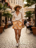 Yanet Collection Shop woman wearing a white linen shirt, orange palm print shorts, straw hat, and white sneakers.