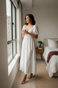 Yanet Collection Shop white maxi dress with puff sleeves, v-neckline, and lace hem worn by a woman by a window.